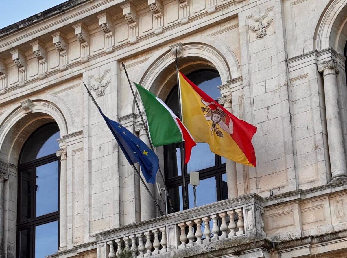 Facciata istituzionale con le bandiere dell’Unione Europea, dell’Italia e della Sicilia, simbolo di identità locale e supporto multilingue per aziende siciliane.